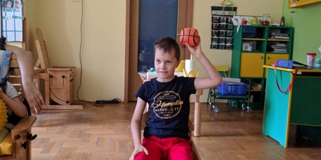 Powiększ grafikę: chłopiec siedzi w lewej ręce trzyma pomarańczową piłkę