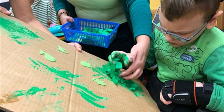 Powiększ grafikę: chłopczyk w zielonej koszulce przykleja do szarego tła zielona bibułę.