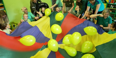 Powiększ grafikę: dzieci i dorośli zielono zebrani wokół spadochronu z zielonymi balonami