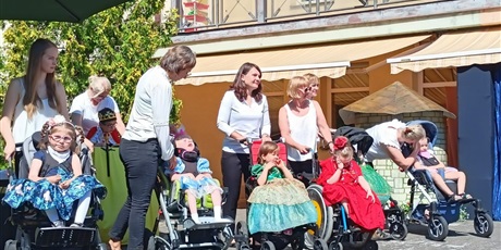 Powiększ grafikę: Na scenie stoją dziewczynki ubrane w książęce suknie, dzieci siedzą na wózkach inwalidzkich.