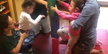 Powiększ grafikę: Dwie dziewczynki i trzech nauczycieli. Dziewczynka w różowej bluzie jest podtrzymywana przez nauczycielkę w blizce w paski i buduje wieżę z klocków. Dziewczynka  wyciąga rękę w kierunku