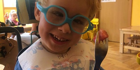 Powiększ grafikę: Uśmiechnięty chłopiec w niebieskich okularach ze śliniakiem siedzi i trzyma w dłoni widelec z nabitym kawałkiem arbuza. 