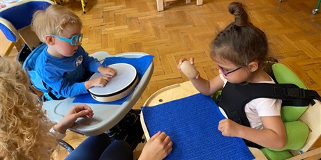 Powiększ grafikę: Chłopiec i dziewczynka siedzą na siedziskach ze stolikami. Przed nimi siedzi nauczycielka. Na stoliku chłopca leży bębenek. Dłonie chłopca leżą na bębenku. Dziewczynka trzyma w dłoni drewniane jajo.  
