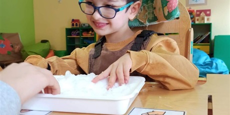 Powiększ grafikę: Chłopiec wkłada ręce do miski pełnej śniegu, zimowe zabawy.