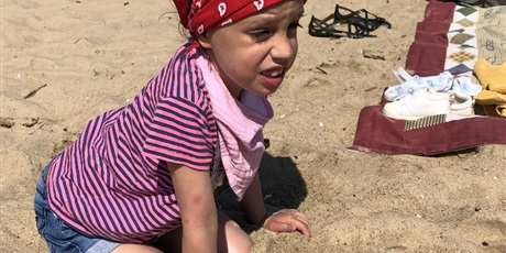 Powiększ grafikę: Na plaży w przysiadzie siedzi dziewczynka w bluzce w paski i czerwonej chustce na  głowie. Dziewczynka jest zmarszczona i krzywi się od słońca 