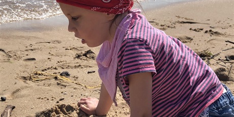 Powiększ grafikę: Na piasku nad brzegiem morza w przysiadzie siedzi dziewczynka w bluzce w paski i czerwonej chustce na  głowie . 