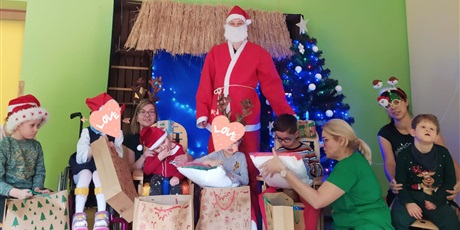 Powiększ grafikę: sześcioro dzieci siedzi przy choince z prezentami, z nimi jest Mikołaj i trzy panie