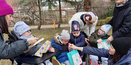 Powiększ grafikę: Dzieci i dorośli w przedszkolnym ogrodzie oglądają prezenty ukryte w papierowych wielkanocnych torebkach. Jedna z dziewczynek i dwóch chłopców siedzi na wózku. 