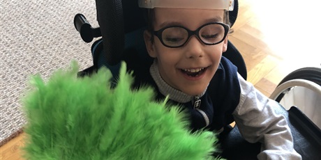 Powiększ grafikę:  Uśmiechnięty chłopiec w okularach siedzi w wózku z głową zabezpieczoną headpodem . Patrzy w kierunku zielonego wachlarza z piór który jest z jego prawej strony. 