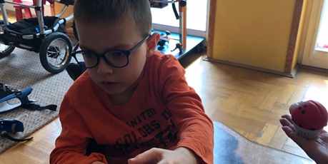 Powiększ grafikę: Chłopiec w okularach, pomarańczowej bluzce siedzi w wózku z blatem. Wyciąga lewą dłoń i próbuje chwycić zieloną, gumową  żabkę trzymaną przed nim. 