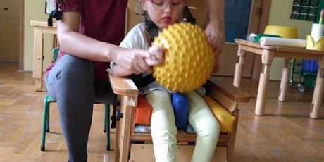 Powiększ grafikę: dziewczynka siedzi na krześle trzyma obiema rękami piłkę z pomocą dorosłego