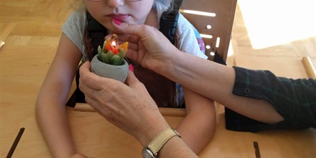Powiększ grafikę: dziewczynka siedzi próbuje dmuchnąć na świeczkę, logopeda pomaga