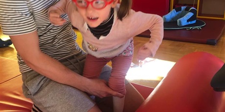 Powiększ grafikę: Dziewczynka w czerwonych okularach stoi przy drabince. Za nią na taborecie siedzi fizjoterapeuta który podtrzymuje i blokuje ją w kolanach. 