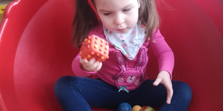 Powiększ grafikę:  W czerwonym, plastikowym stożku siedzi dziewczynka z kitkami. Między nogami ma gumowe zabawki. W prawej dłoni trzyma gumową kostkę i patrzy na nią. 