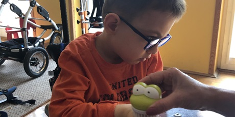 Powiększ grafikę: Chłopiec w okularach, siedzi na wózku z przezroczystym blatem a dorosłe dłonie z zieloną żabką- szczoteczką dotykają jego prawej dłoni. Chłopiec odwraca głowę w drugą stronę.